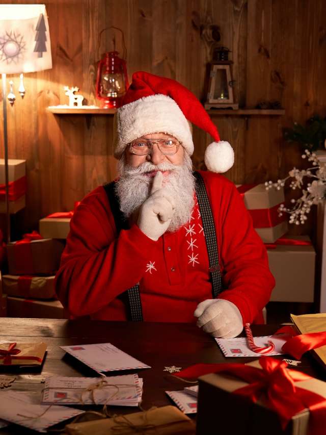 Santa Claus showing shh secret gesture on Christmas eve sitting at home table.