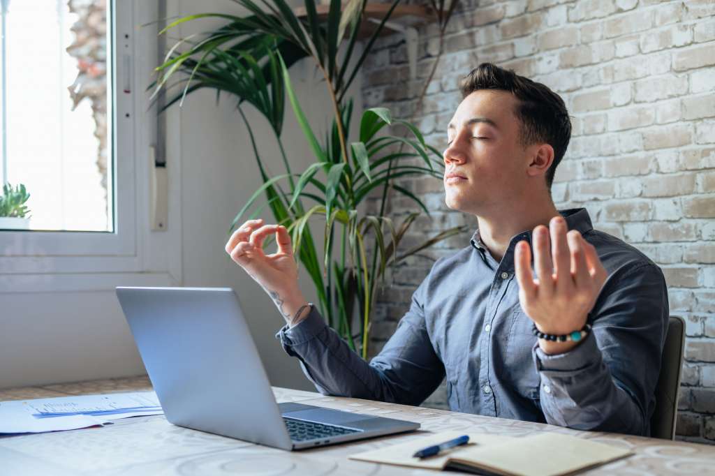 Serene office male employee sit at desk relaxing doing yoga, practice meditation to reduce stress