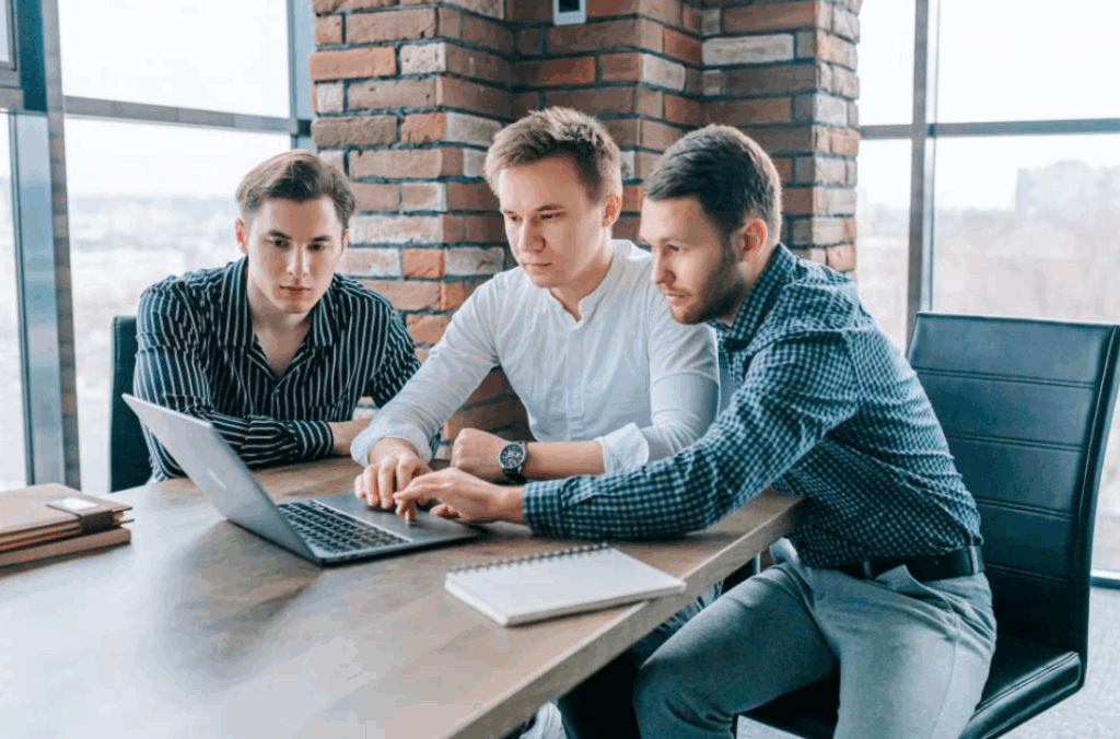 Three guys working on their laptops, with a trainer providing support and feedback
