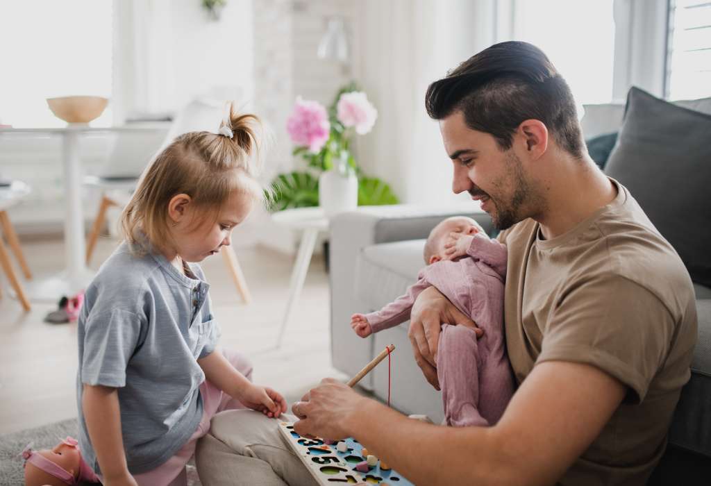 Happy young man taking care of his newborn baby and little daughter indoors at home, paternity leave
